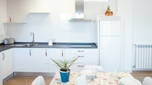 A modern kitchen with white cabinets, a stainless steel range hood, a built-in oven, and a white refrigerator. There is a dining table with chairs and a potted plant. - Pico Rubia, two bedroom apartment, living room, kitchen and bathroom (Ampuero)