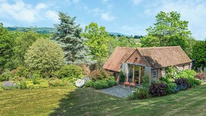 A small brick house with a sloped roof, a glass door entrance, and a garden with various plants and a bench. - The Cottage High Wykehurst End (Ewhurst, near Cranleigh)