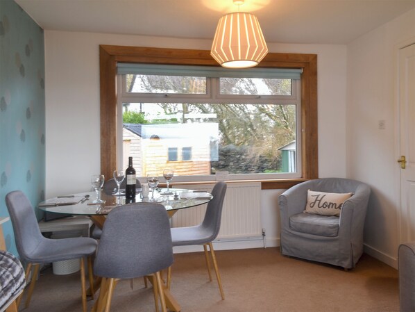Un coin repas avec une table dressée pour deux personnes, une chaise, un canapé agrémenté d’un coussin orné du mot « Accueil », et une fenêtre donnant sur une maison et des arbres.
