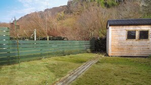 A wooden cabin with a metal roof, surrounded by a green fence and a stone wall. - 2 bedroom accommodation in Ellenabeich, near Oban (Ellenabeich, near Oban)