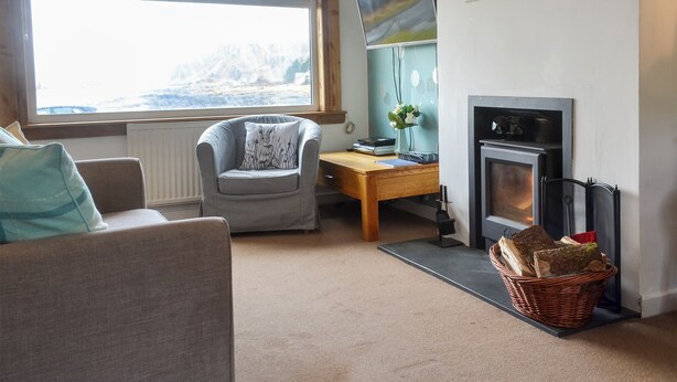 A cozy living room with a fireplace, a comfortable armchair, a wooden desk, and a large window offering a view of the outdoors. - 2 bedroom accommodation in Ellenabeich, near Oban (Ellenabeich, near Oban)