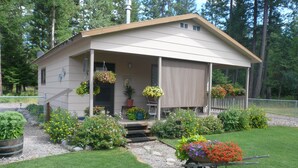A small, single-story house with a porch, surrounded by a well-maintained garden and a wheelbarrow of flowers. - GUEST CABIN IN THE WOODS (Bigfork)