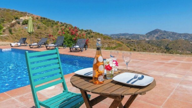 Mesa à beira da piscina com uma garrafa, copos e um prato, cercada por espreguiçadeiras e vista para colinas.