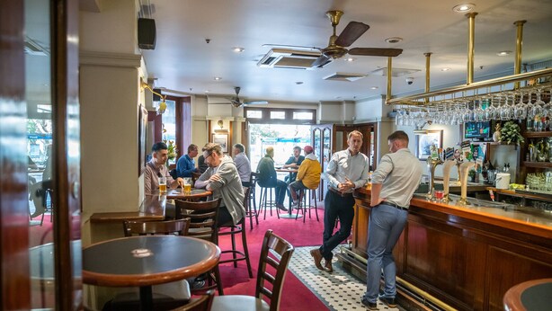 A bar area with tables and chairs, a counter with a bartender, and a ceiling fan. - The Occidental Hotel (Sydney)