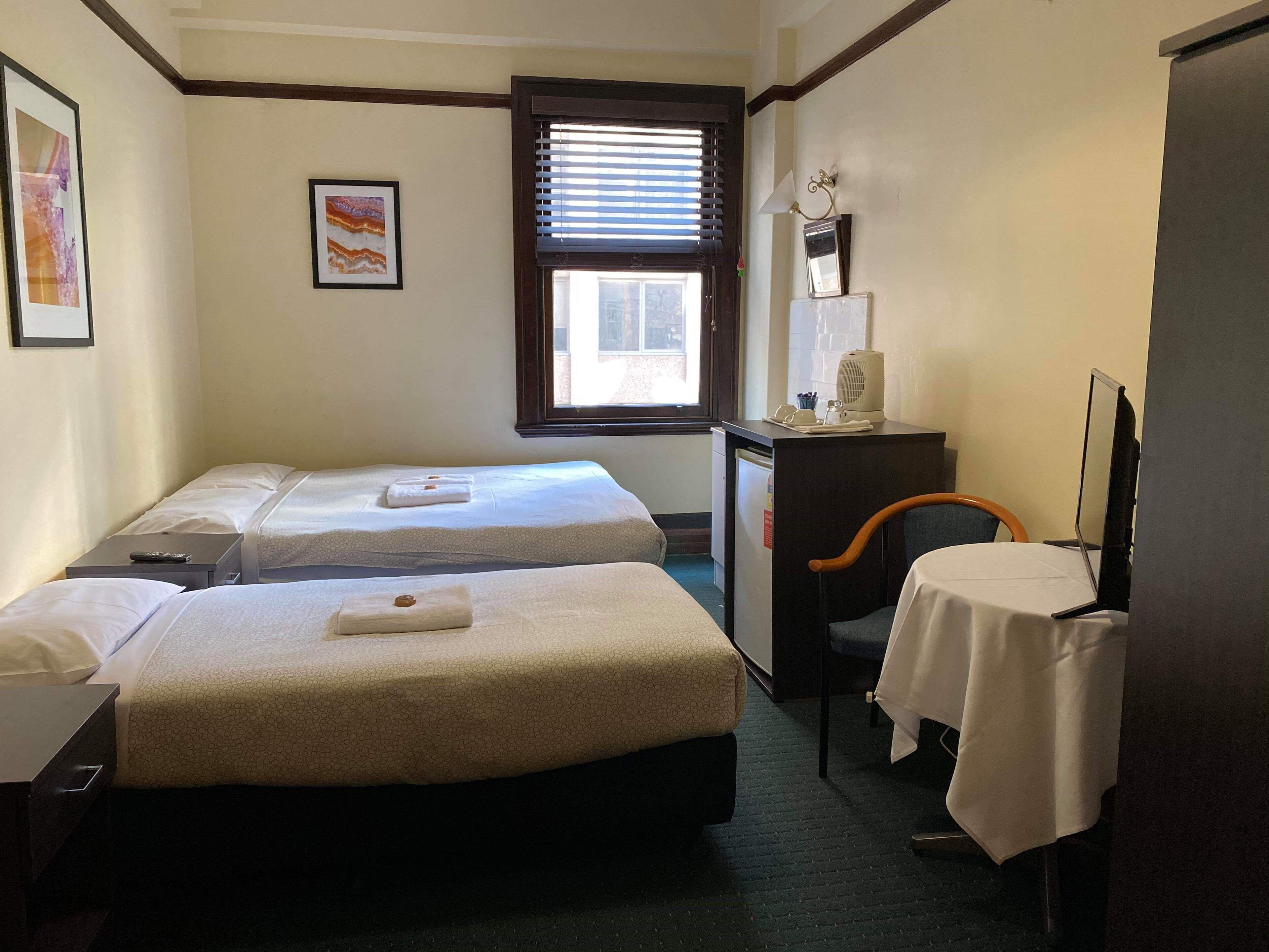 Une chambre d’hôtel avec deux lits, un bureau avec une chaise, un petit réfrigérateur et une fenêtre avec des stores.