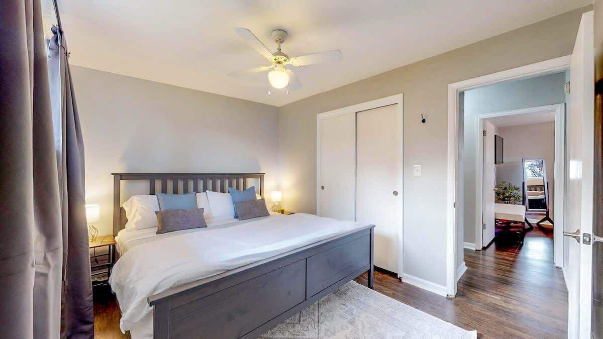 Une chambre à coucher comprenant un lit, des tables de chevet, un ventilateur de plafond et une porte donnant sur une autre pièce.