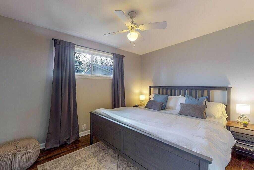 A bedroom with a bed, curtains, a ceiling fan, and a nightstand with a lamp.