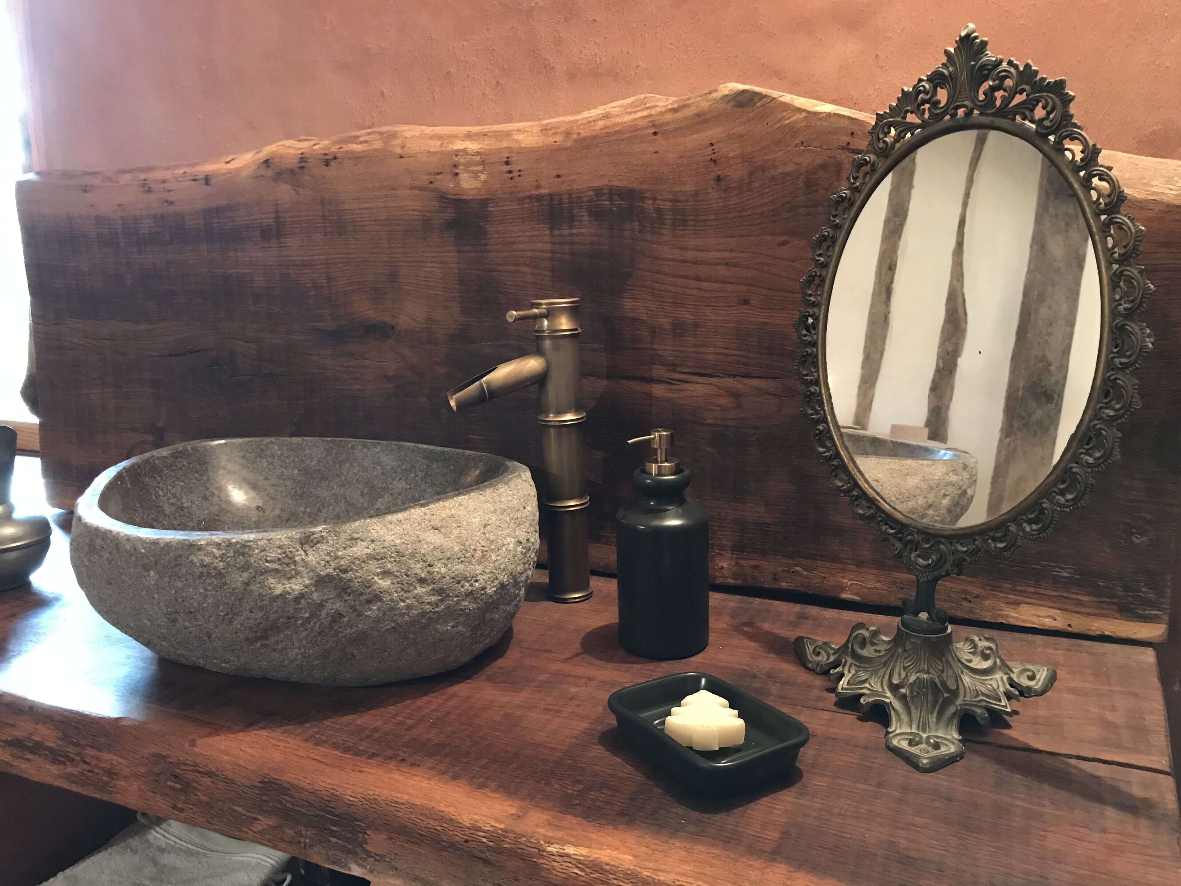 A stone sink, a brass faucet, a black soap dispenser, a small black soap dish, and an ornate mirror on a wooden surface.