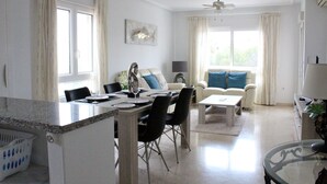 A spacious living area with a dining table, chairs, a sofa, and a TV. There are ceiling fans, a window with curtains, and a plant on a table. - 2 Bed 2 Bath South Facing Apt, Quiet But Near Beaches and Amenities (Orihuela)