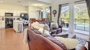 A spacious living area with a leather sofa, dining table, and chairs, a kitchen with modern appliances, and large windows offering a view of the outdoors. - Gathering Resort Pool/Spa Home-Mins away to the PARKS (Kissimmee)