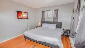 Une chambre à coucher avec un grand lit, une table de chevet, un téléviseur fixé au mur et une fenêtre avec des rideaux.