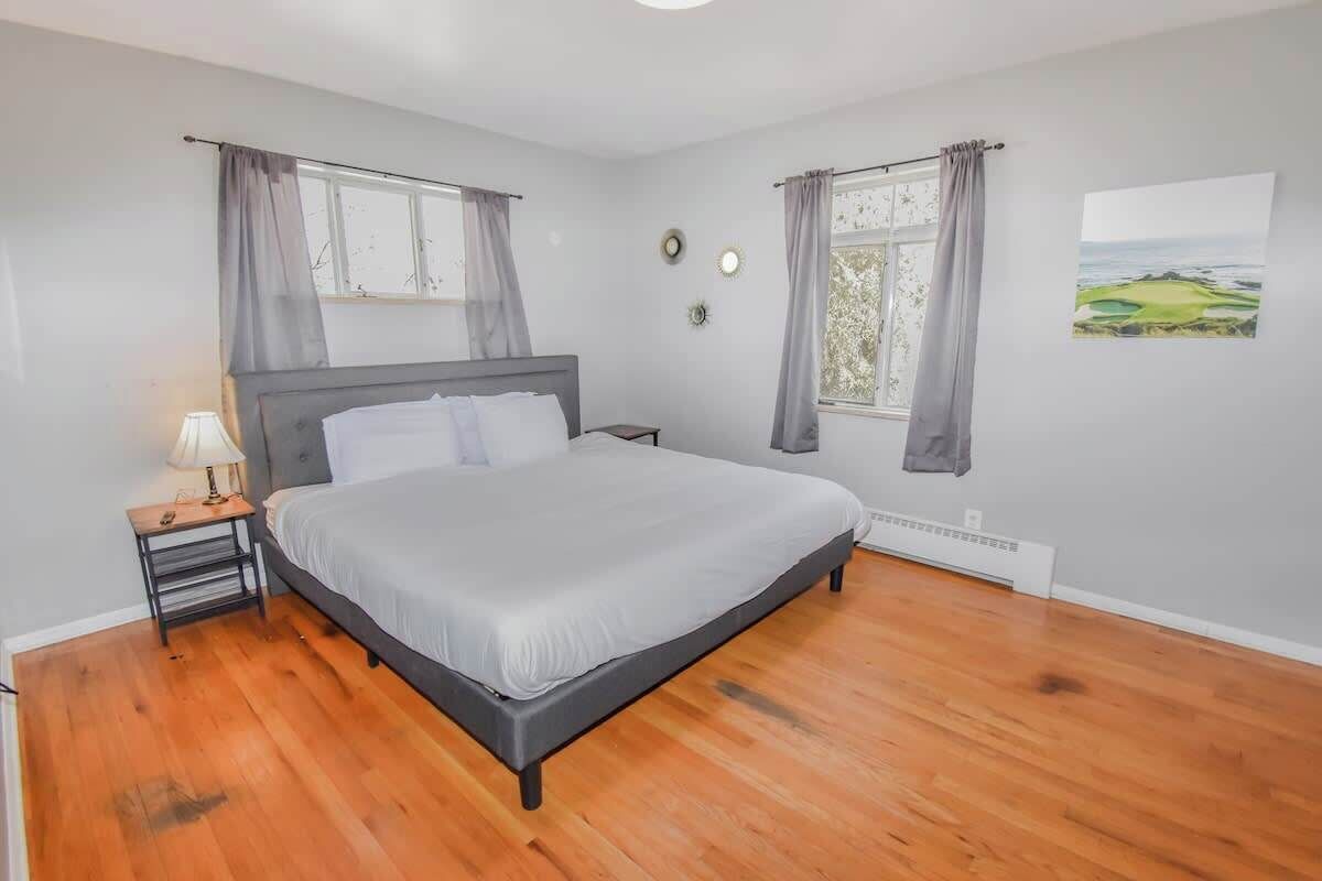 Un dormitorio con una cama, una mesita de noche, una lámpara, dos ventanas con cortinas y un termostato.
