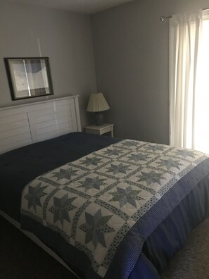 Une chambre à coucher avec un lit, une table de chevet avec une lampe, un tableau encadré au mur et une fenêtre avec des rideaux.
