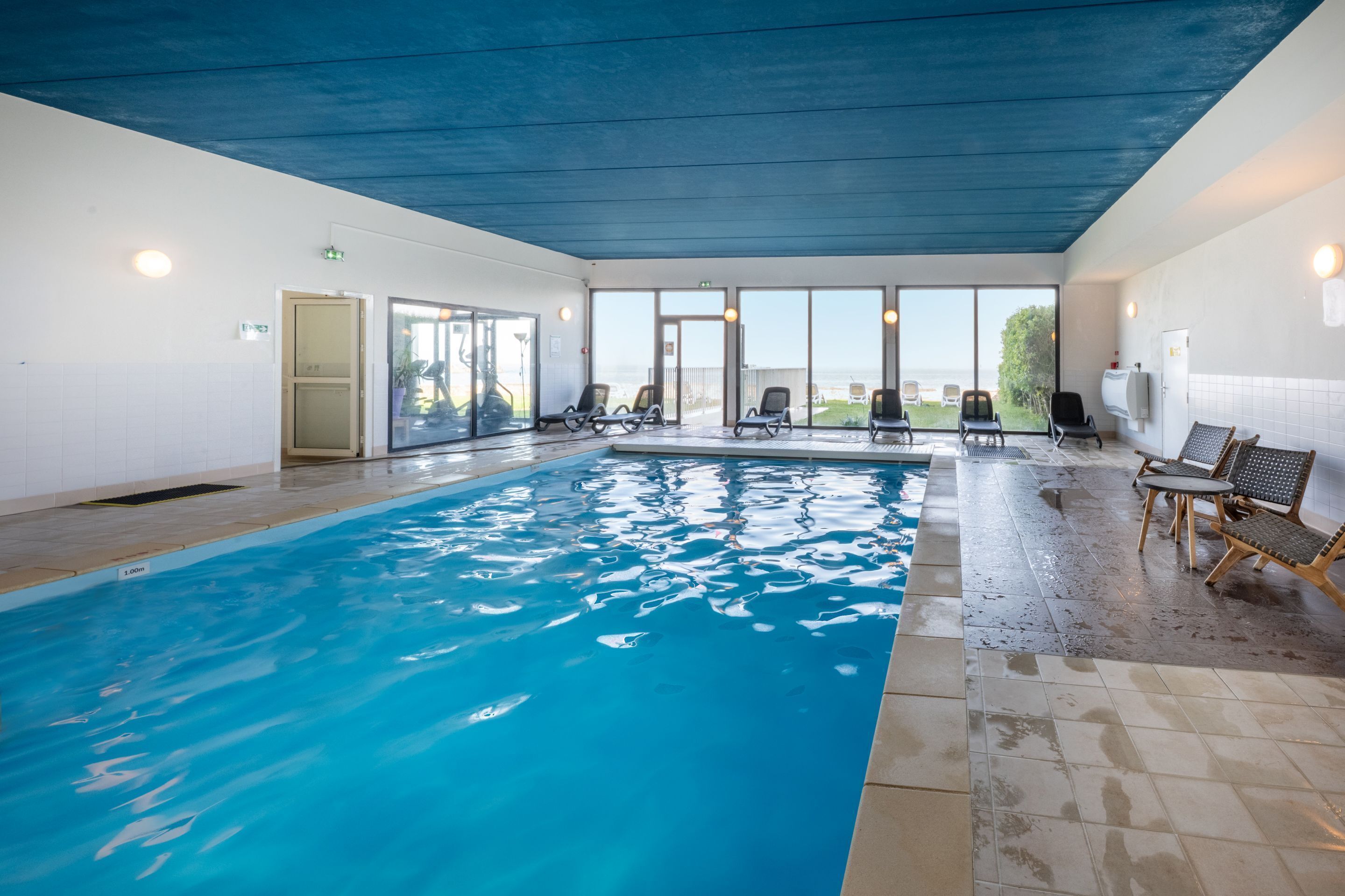 Une piscine intérieure à l’eau claire, entourée de chaises longues et offrant une vue sur la mer grâce à de grandes fenêtres.