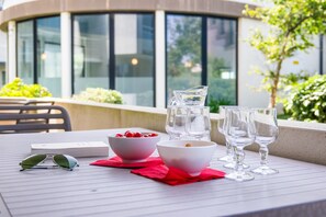 A table with a bowl of cherries, a pitcher, and wine glasses, set on a terrace with a view of a building and greenery.