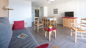 Ein modernes Wohnzimmer mit einem grauen Sofa, roten Kissen, einem Essbereich mit Holztischen und -stühlen, einem Fernseher auf einem roten Schrank und einem Flur, der zum Küchenbereich führt.