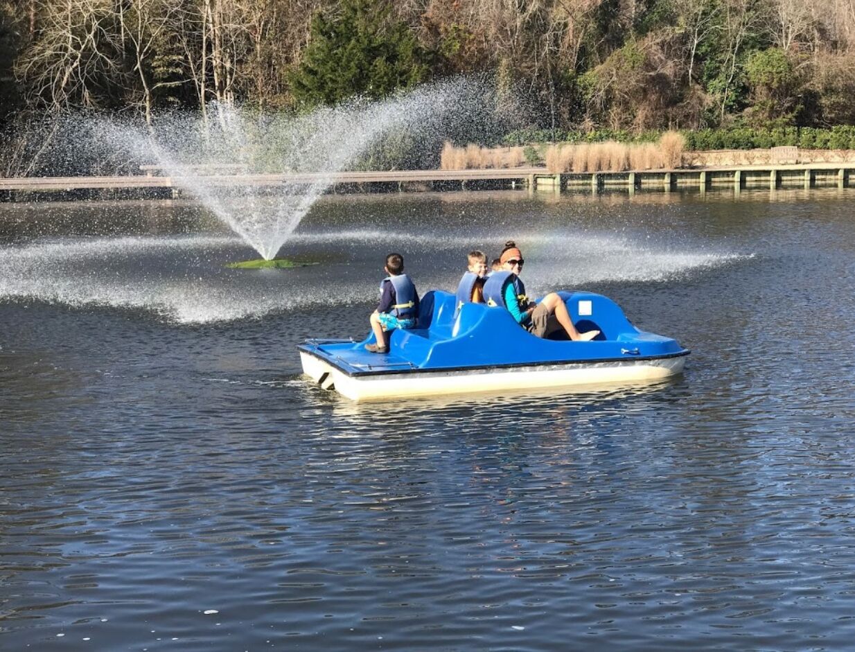 A blue boat with passengers on a calm lake, surrounded by trees and a fountain.