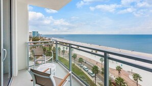 A balcony with a view of the beach and ocean, a table, and chairs.