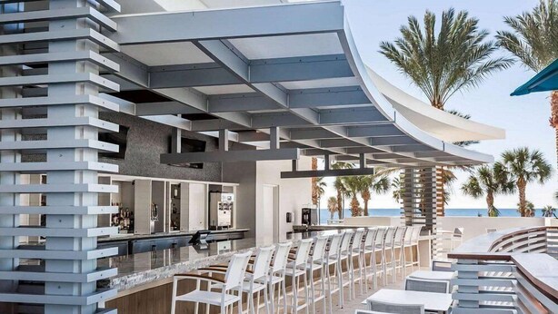 A modern outdoor bar area with a curved ceiling, white bar stools, and a view of the ocean.