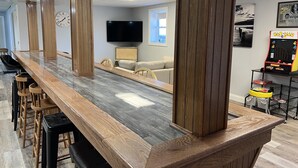 Una barra de madera larga con taburetes altos, un televisor de pantalla plana y una máquina arcade de Pac-Man en una habitación con un sofá y una ventana.