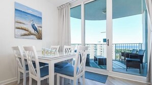 Un comedor con una mesa y sillas blancas, un cuadro de un pájaro en la pared y un balcón con vista al mar.