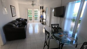 A living room with a black leather sofa, a glass dining table with chairs, a ceiling fan, a TV mounted on the wall, and a door leading to a balcony.