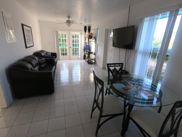 A living room with a black leather sofa, a glass dining table with chairs, a ceiling fan, a TV mounted on the wall, and a door leading to a balcony.
