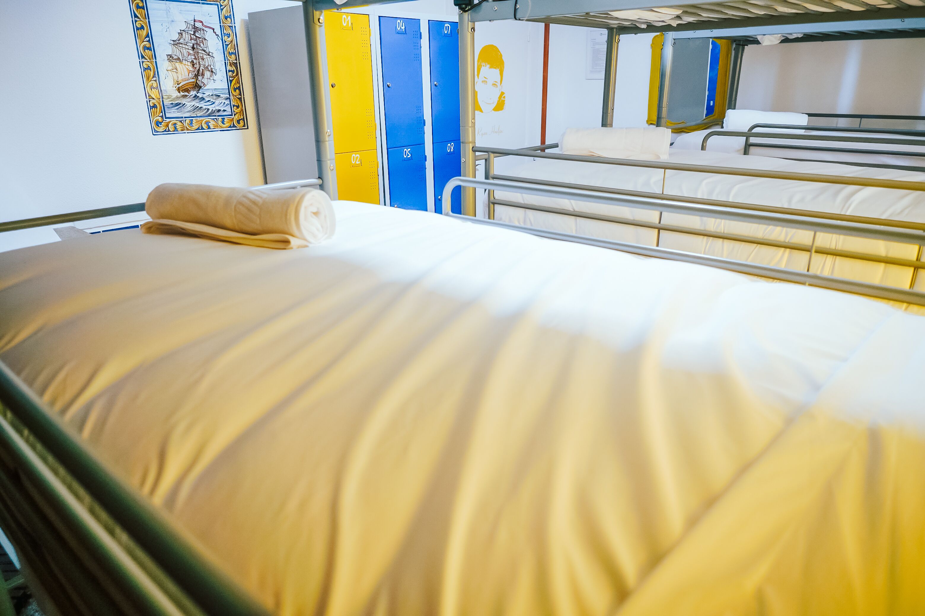 A bunk bed with a neatly made bed and a towel. There are lockers in yellow, blue, and white, and a framed picture on the wall.