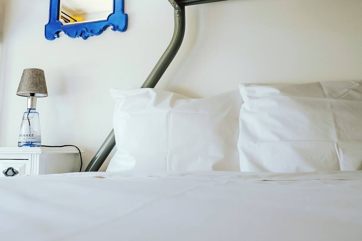 A neatly made bed with white linens, a lamp, and a blue-framed mirror on the wall.