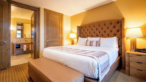 A hotel room with a large bed, a wooden headboard, bedside lamps, a wooden nightstand, and a bathroom visible through an open door.
