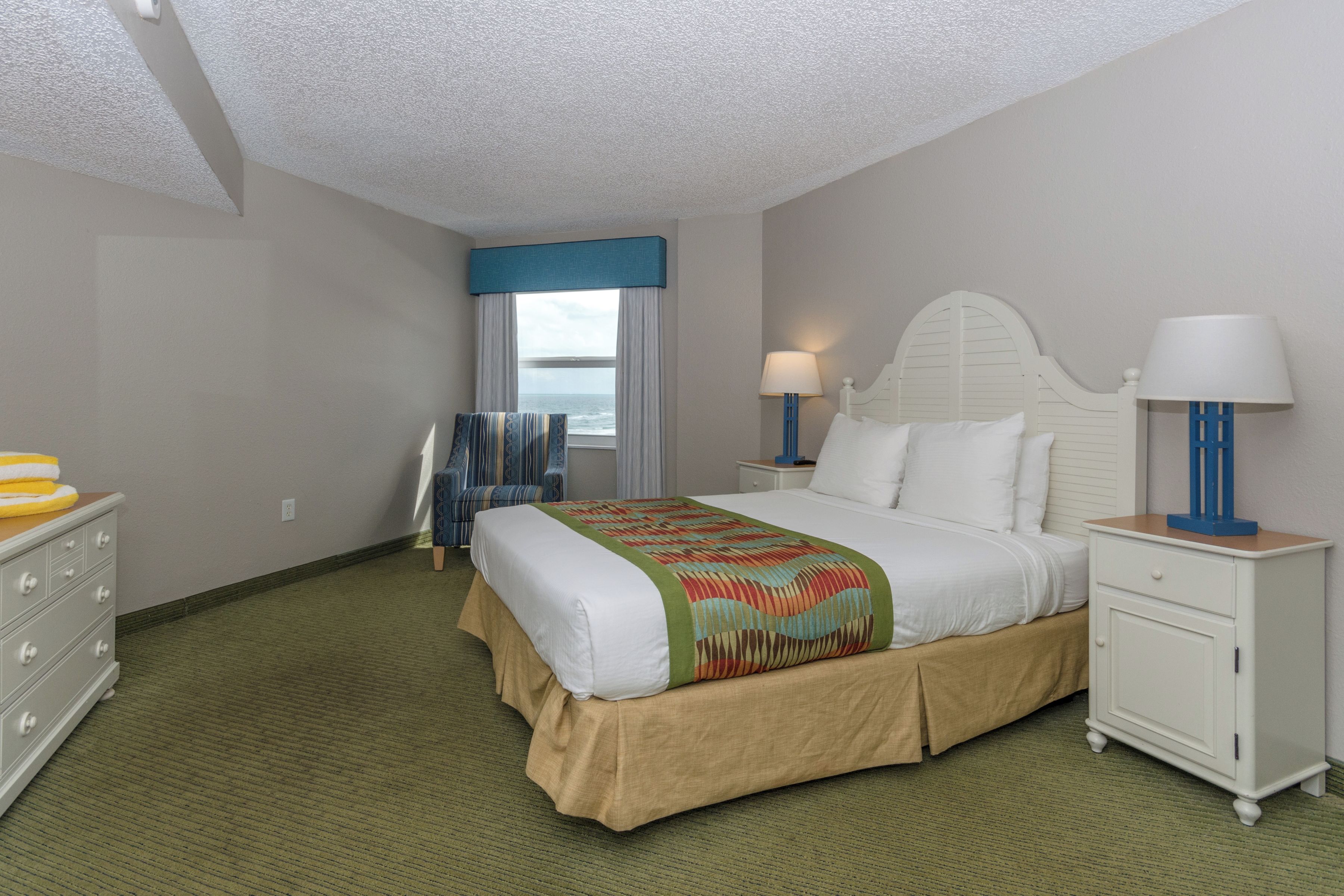 A hotel room with a bed, nightstands, a dresser, and a view of the ocean.