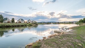 A calm lake with a picnic table and benches on the shore, surrounded by houses and greenery.