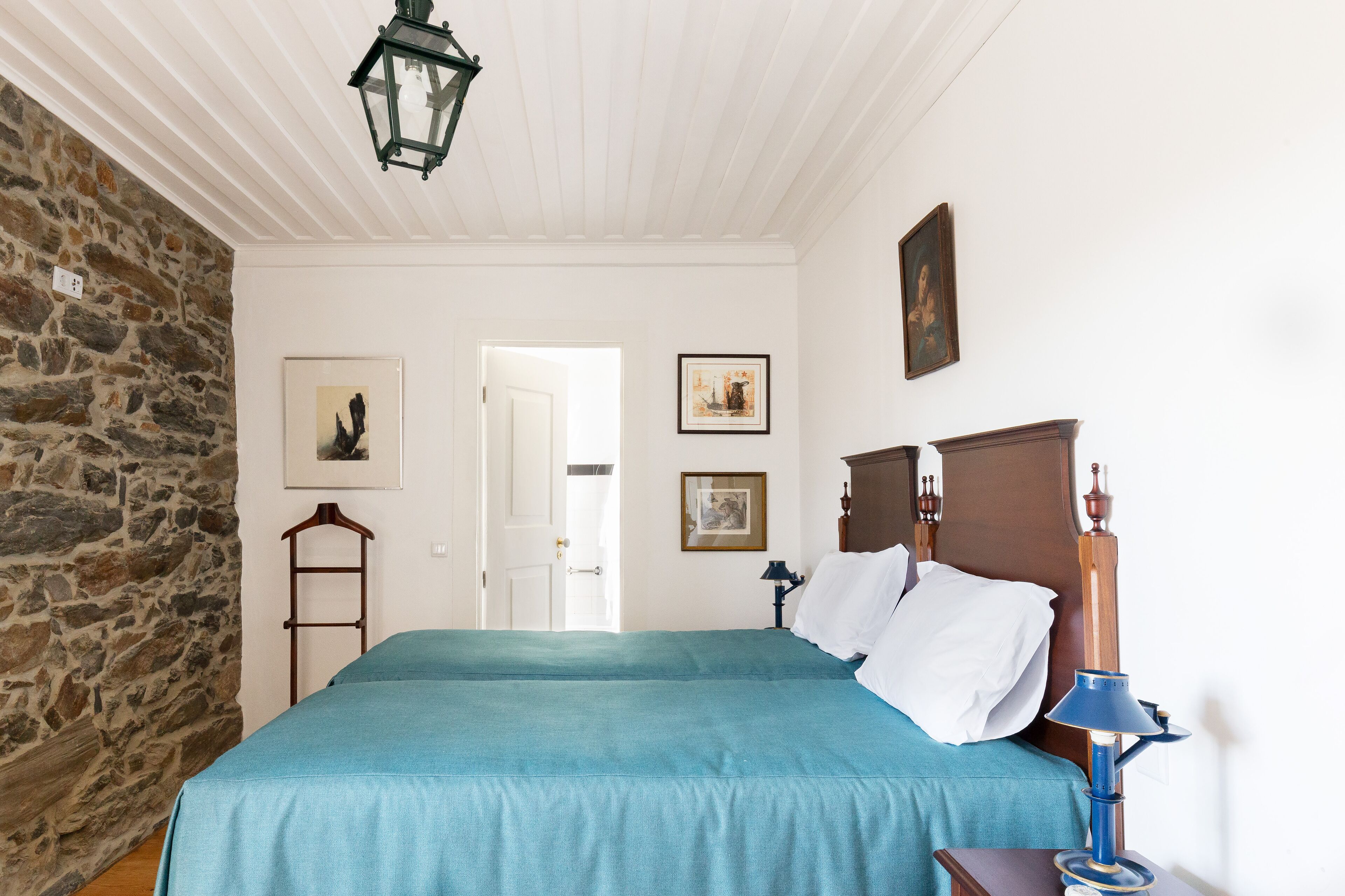 A bedroom with a stone wall, a bed with a blue bedspread, a wooden headboard, and a nightstand with a lamp.
