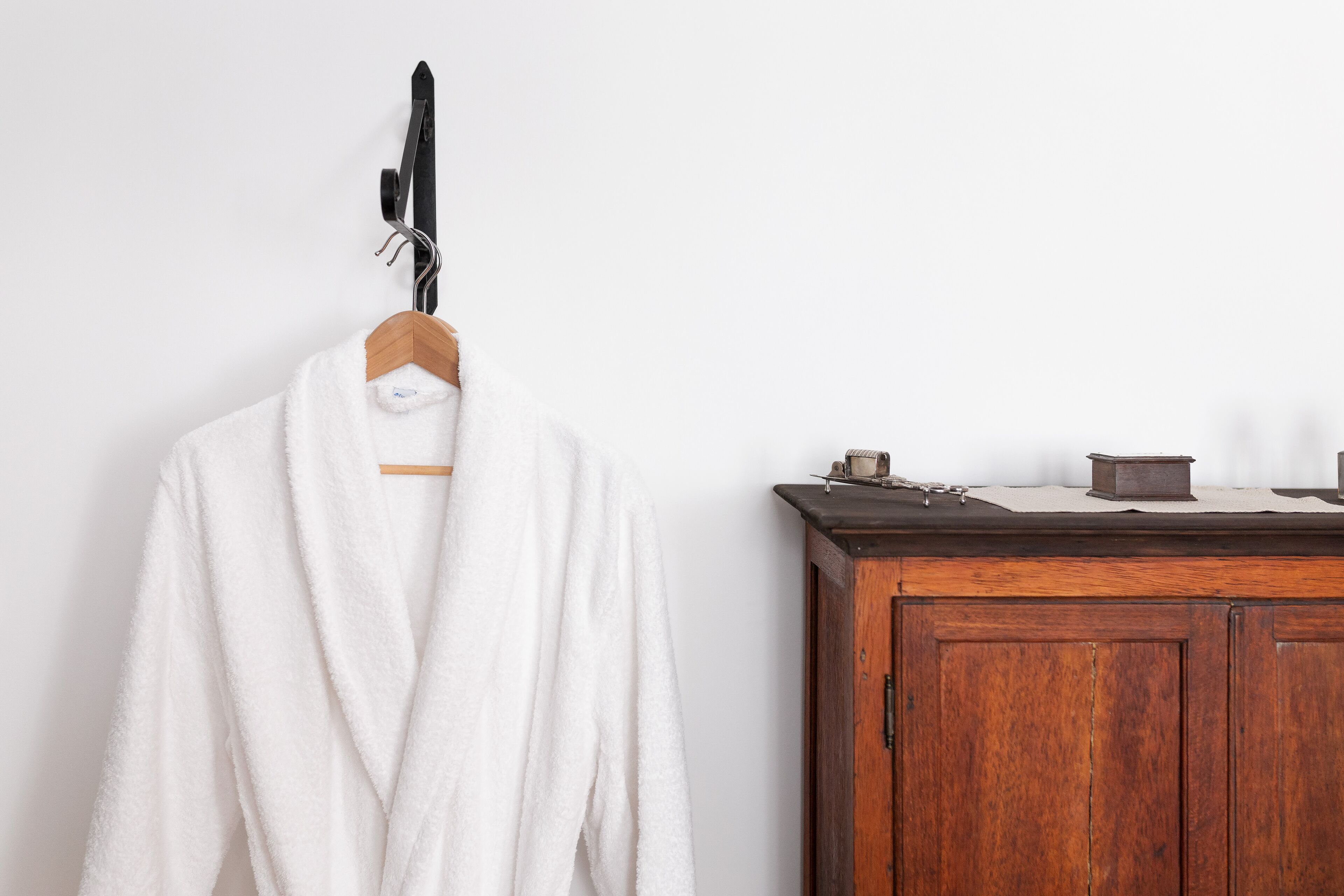 A white bathrobe hanging on a wooden hanger against a white wall, next to a wooden cabinet with a dark countertop.