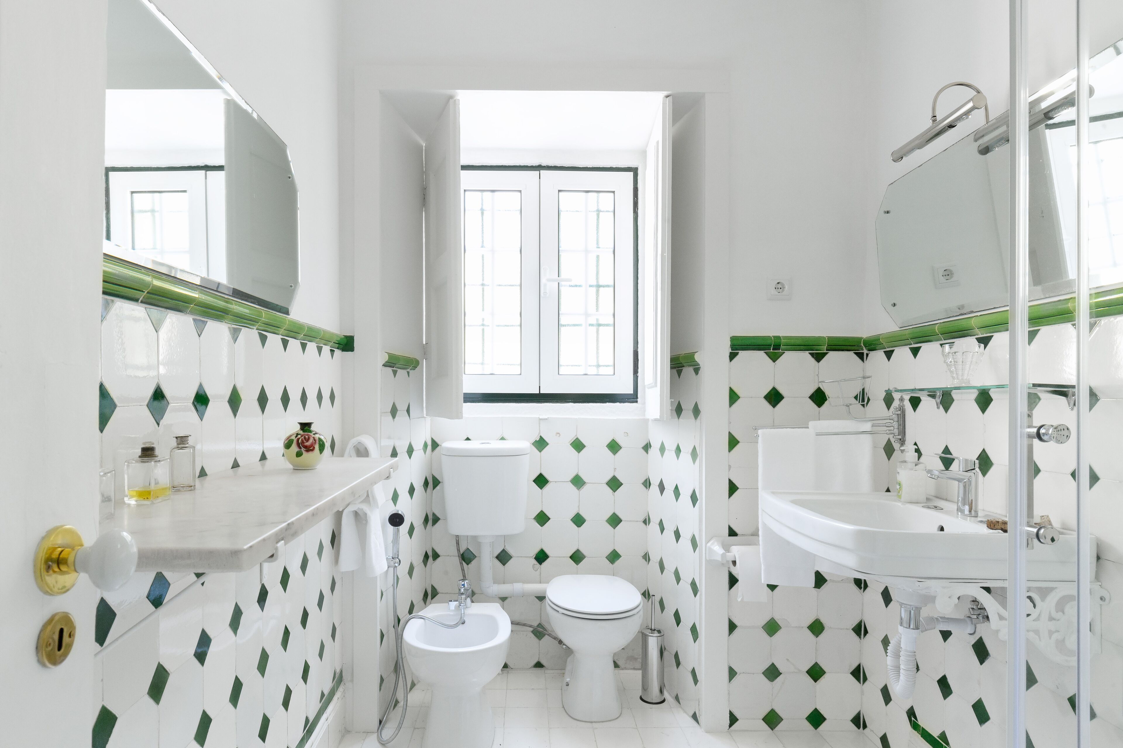 Un baño con inodoro, bidé y lavamanos, con azulejos verdes de diseño de diamantes y una ventana.