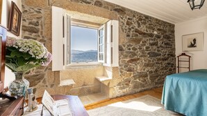 Una habitación con pared de piedra, ventana con contraventanas abiertas, cama con colcha color turquesa y un jarrón con flores.