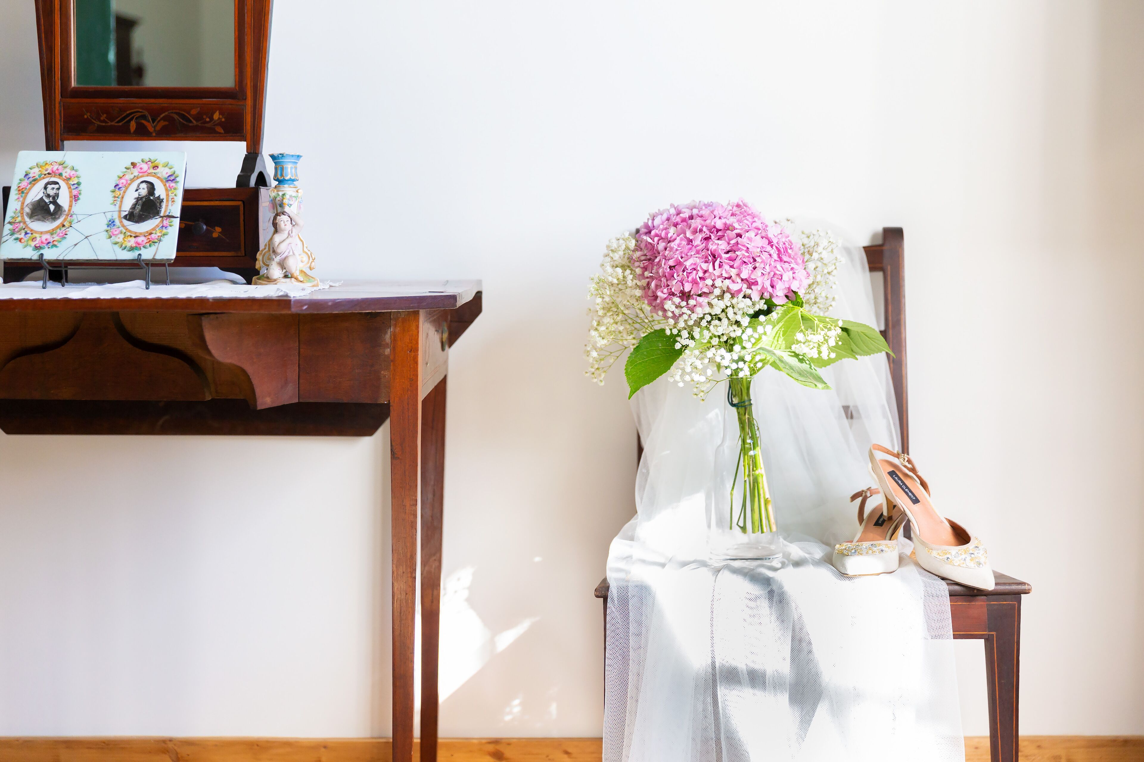 Ein hölzerner Stuhl mit weißem Stoff, eine Vase mit rosa Blumen und ein Paar Schuhe auf einem Tisch.