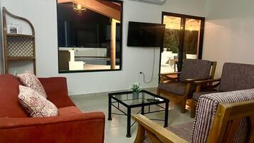 Un salon avec un canapé rouge, une table basse en verre et des fauteuils à rayures. Un téléviseur à écran plat est fixé au mur, et un appareil de climatisation est installé juste au-dessus.