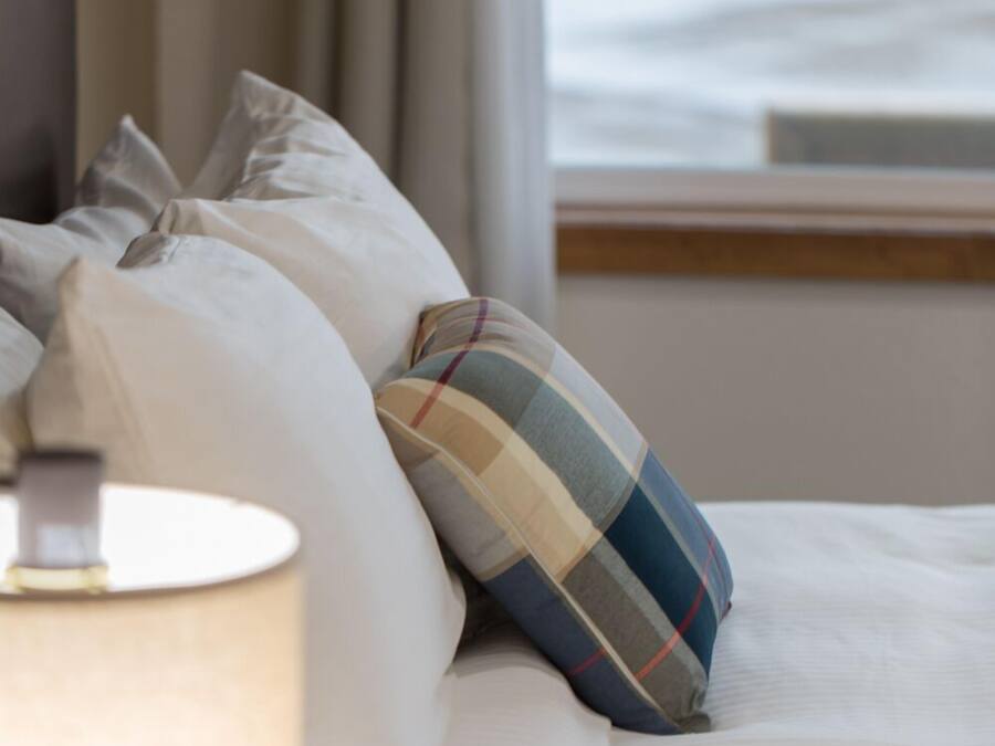 Un letto con biancheria bianca e cuscino a quadri, una lampada sul comodino e una finestra con tende.