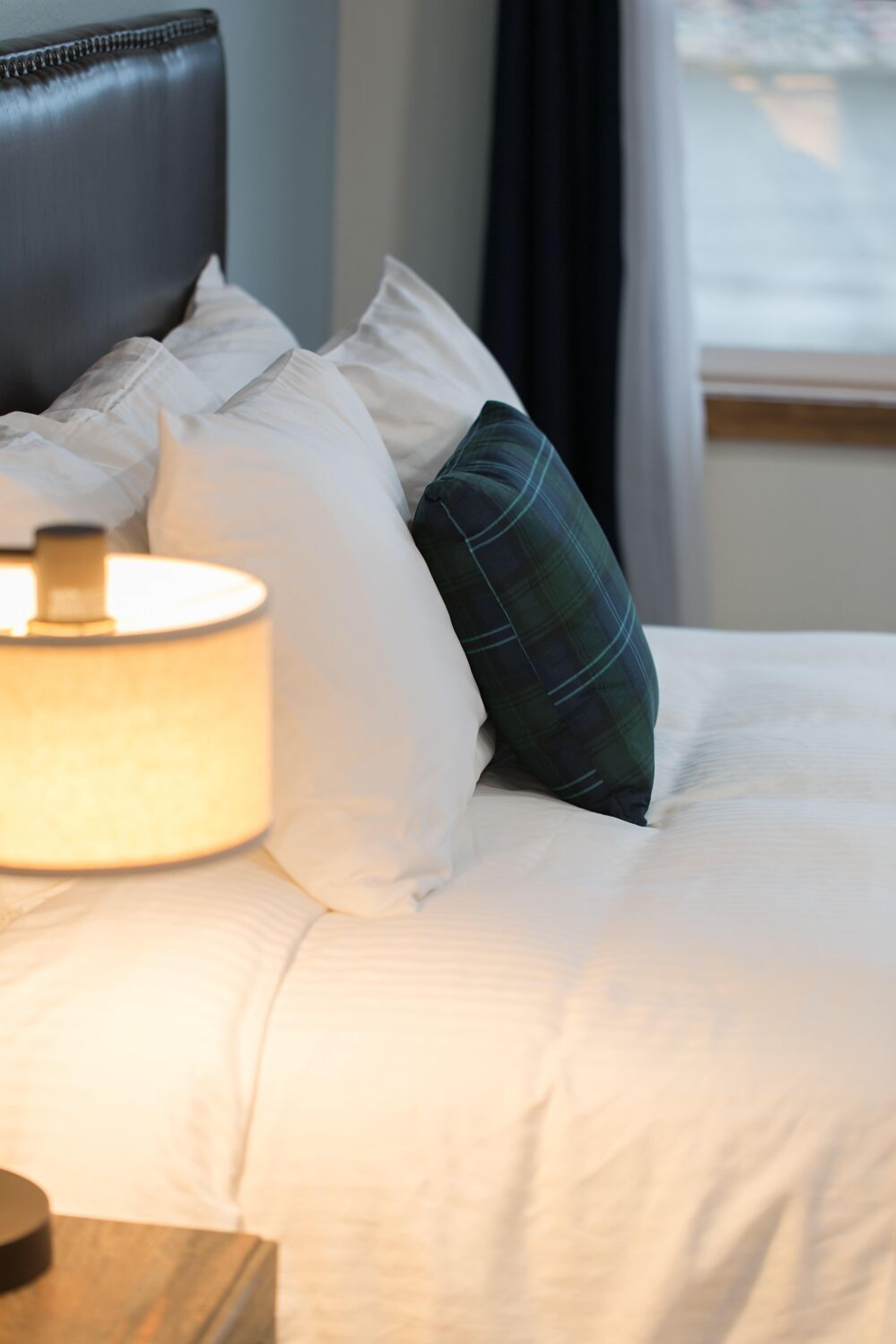 A neatly made bed with white linens and a plaid pillow, a bedside lamp, and a dark headboard.