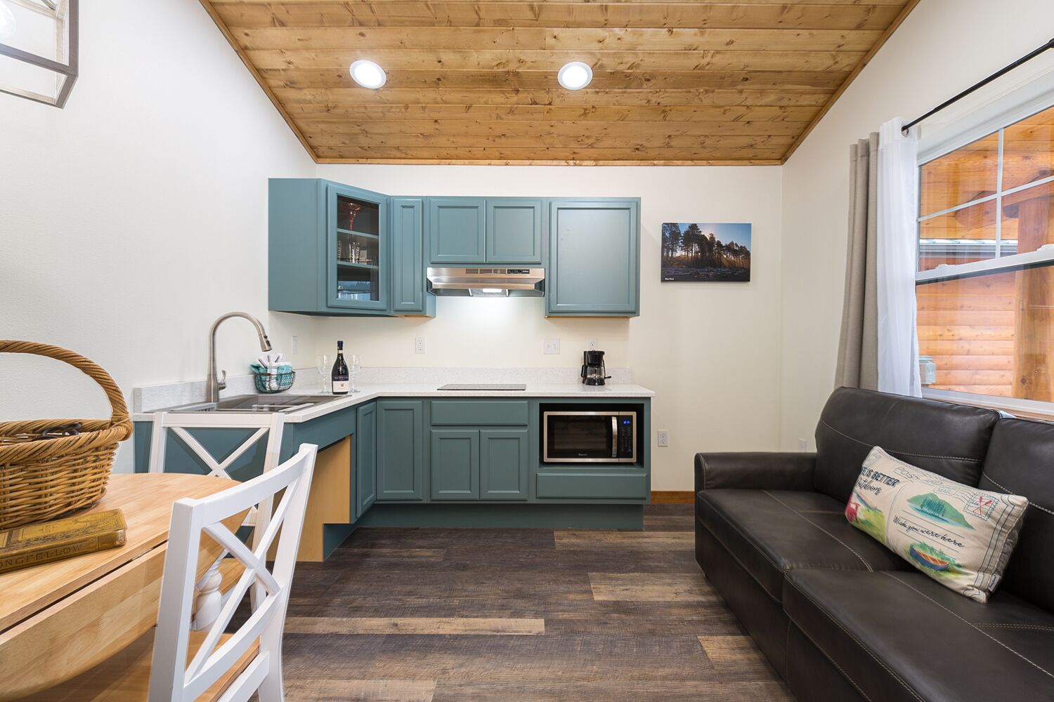 A compact kitchen with teal cabinets, a microwave, and a coffee maker. There is a black leather sofa with a cushion and a window with curtains.