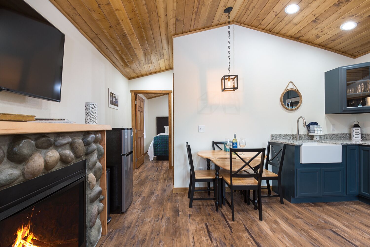 Un cuarto acogedor con chimenea de piedra, techo de madera, una mesa de comedor con sillas, una zona de cocina con armarios azules y un televisor en la pared.
