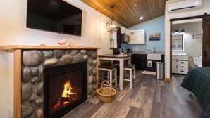 A cozy room with a stone fireplace, a flat-screen TV, a wooden ceiling, and a kitchen area with a sink and cabinets.