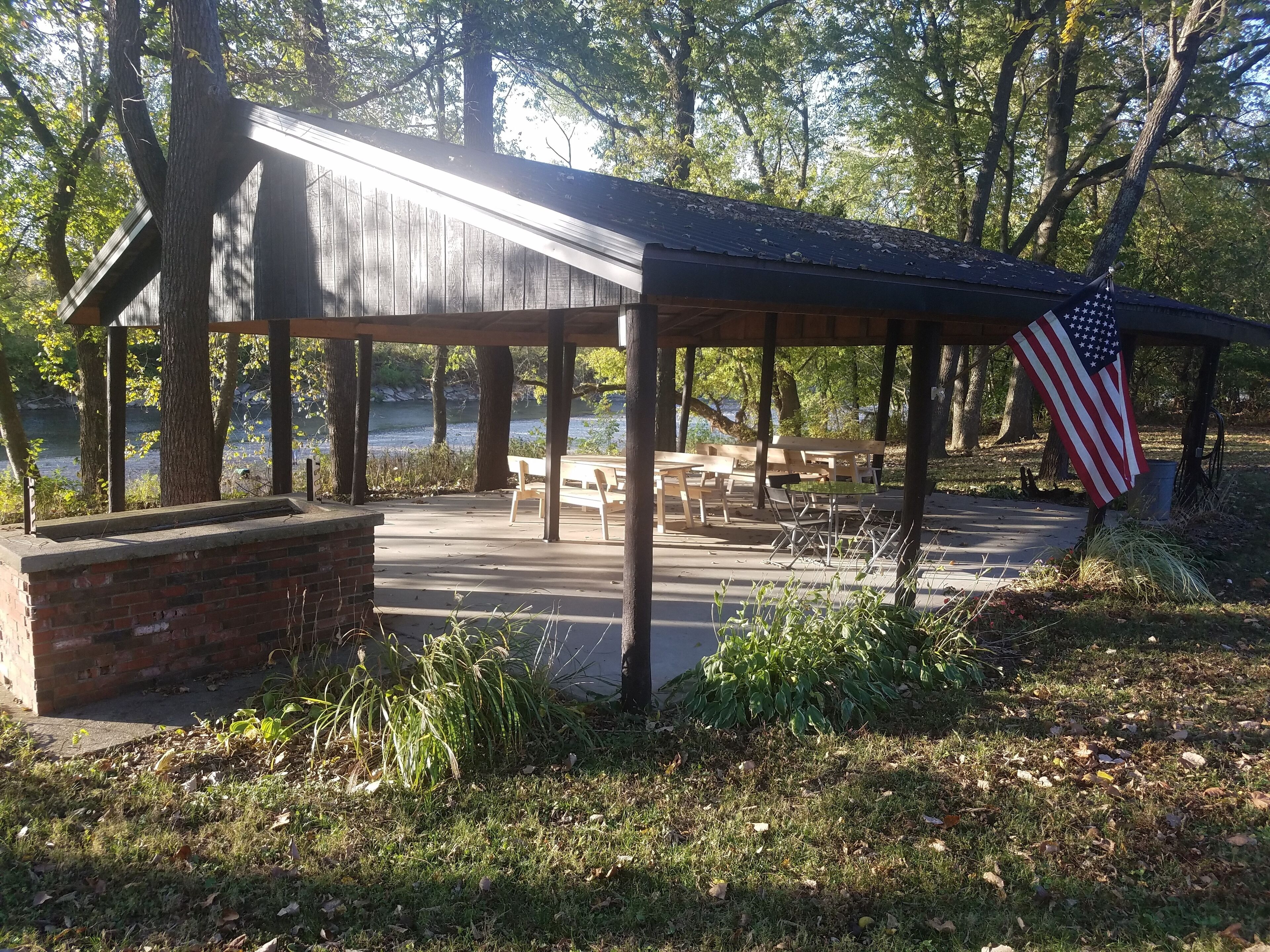 Un espace pique-nique abrité, équipé de bancs et de tables, avec un drapeau américain et une vue sur un lac.