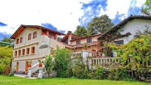 Um edifício de dois andares com fachada branca e telhado de telhas vermelhas, cercado por vegetação exuberante e uma parede de pedra.