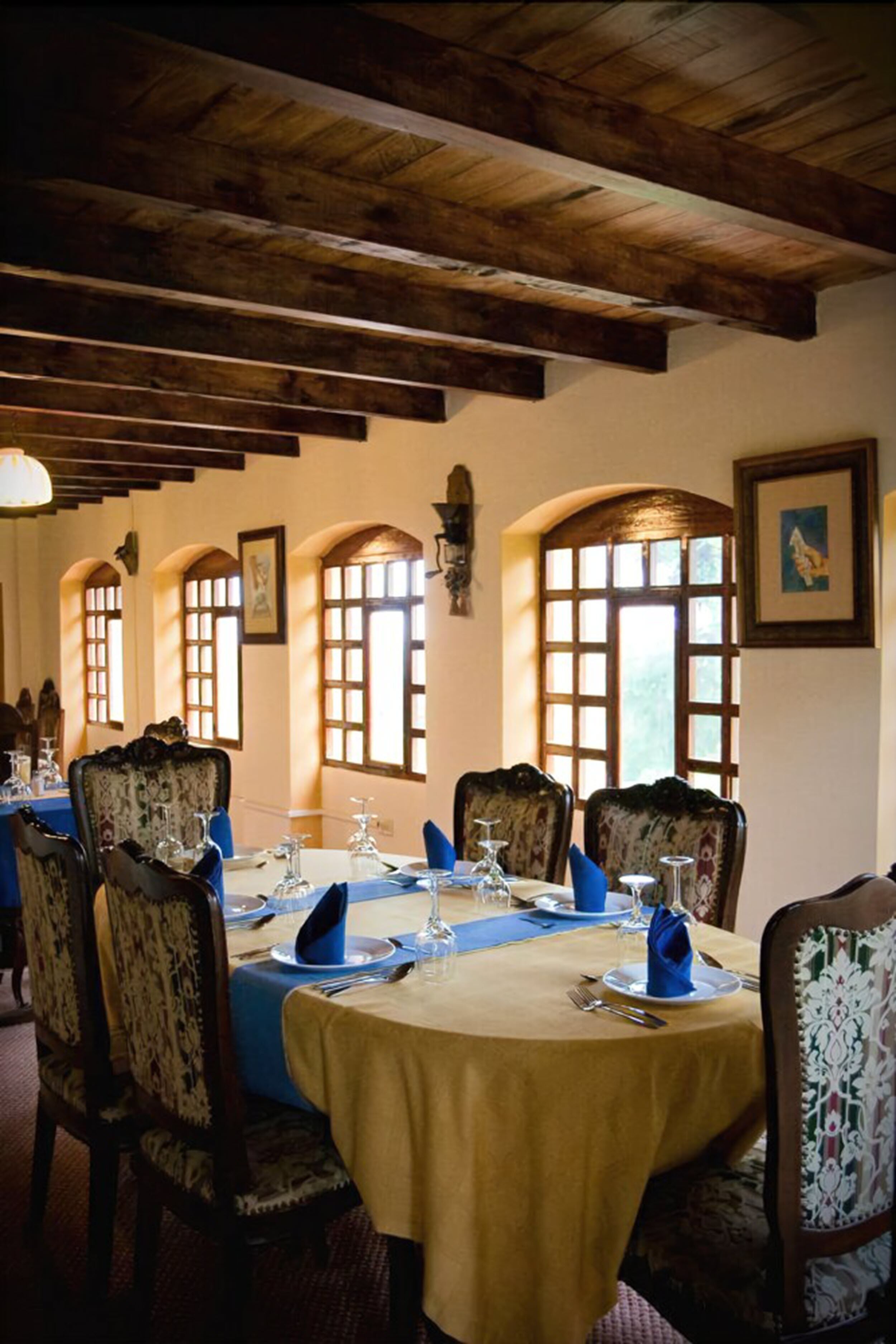 Uma sala de jantar com uma mesa redonda posta para quatro pessoas, vigas de madeira no teto e janelas em arco.