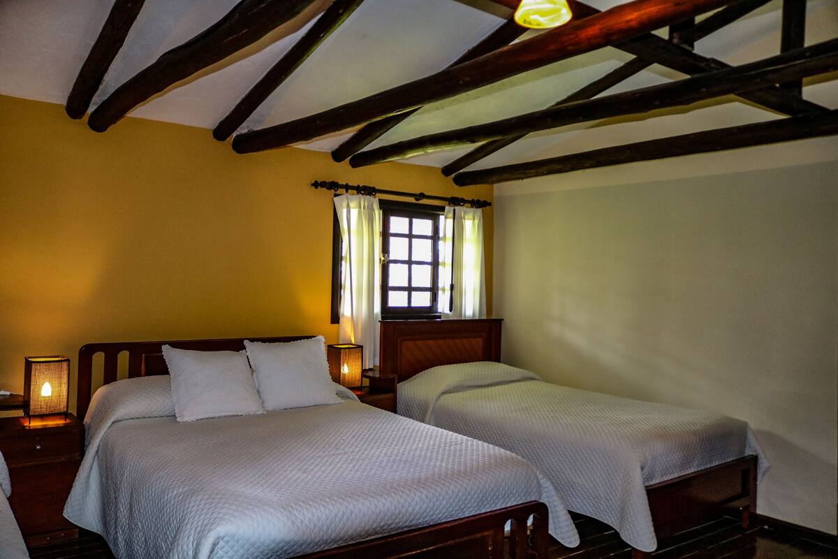 A room with two beds, wooden beams on the ceiling, a window with curtains, and a bedside lamp.