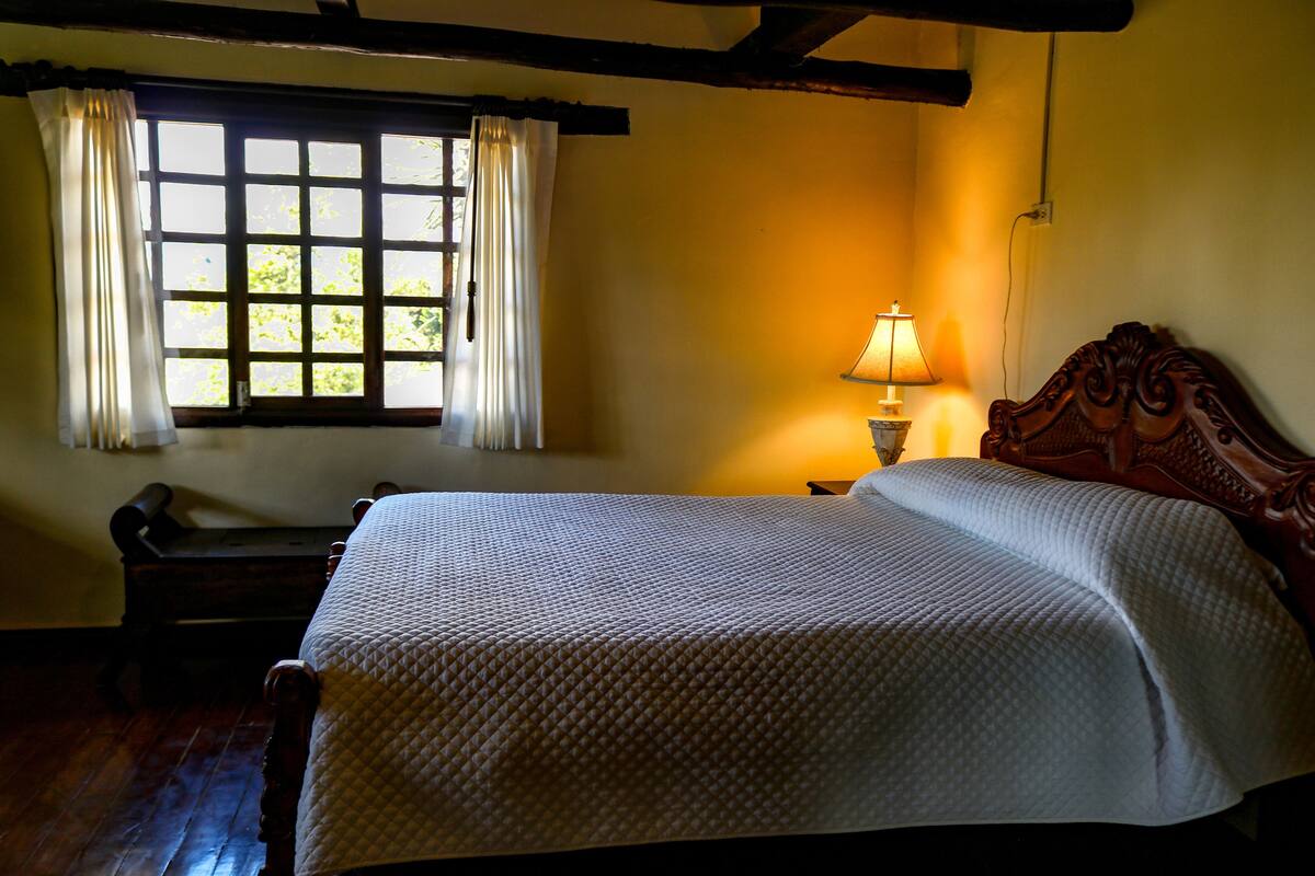 A neatly made bed with a quilt, a window with curtains, and a bedside lamp in a room with wooden beams.