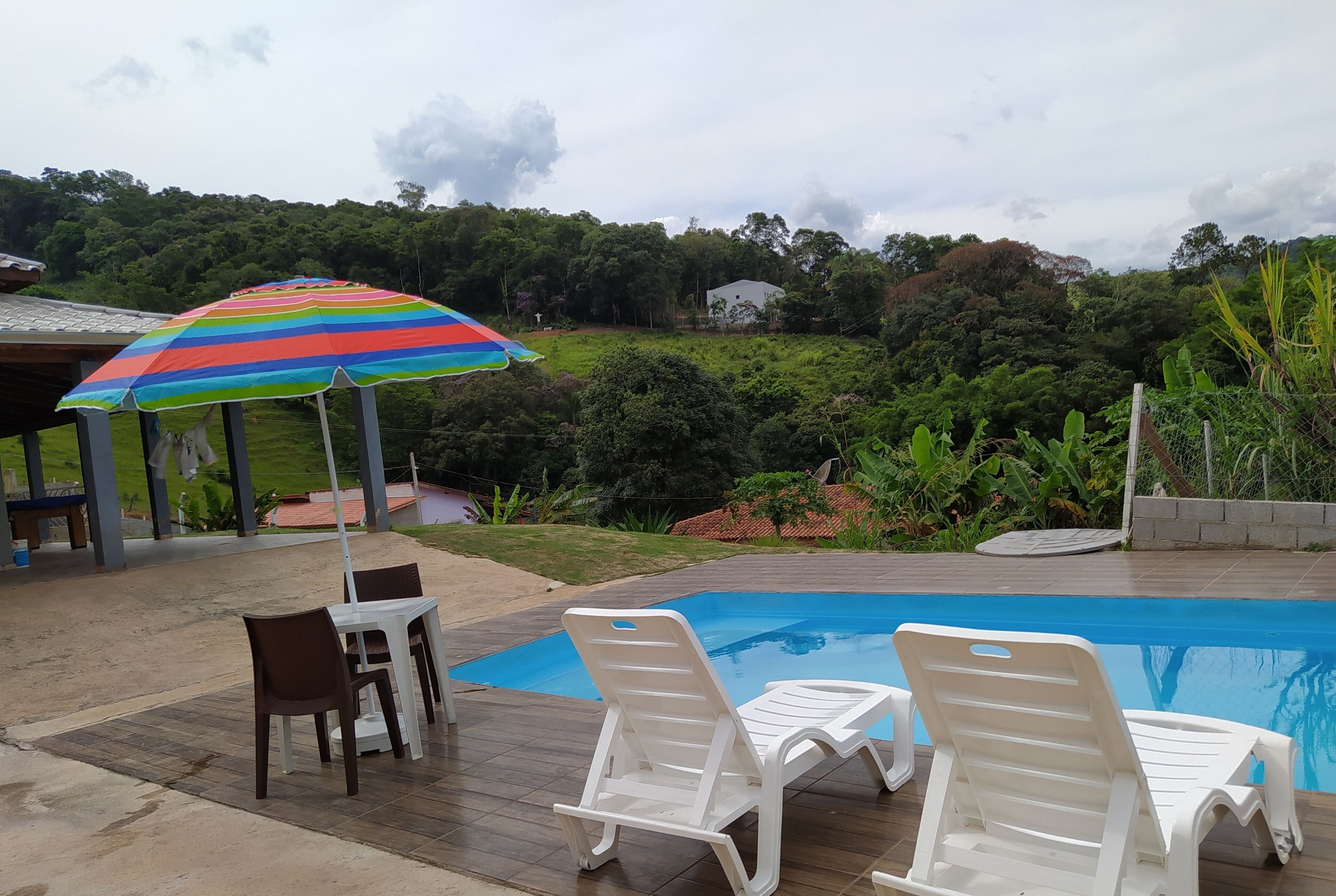 A pool area with lounge chairs and a table, surrounded by greenery and a house in the background.