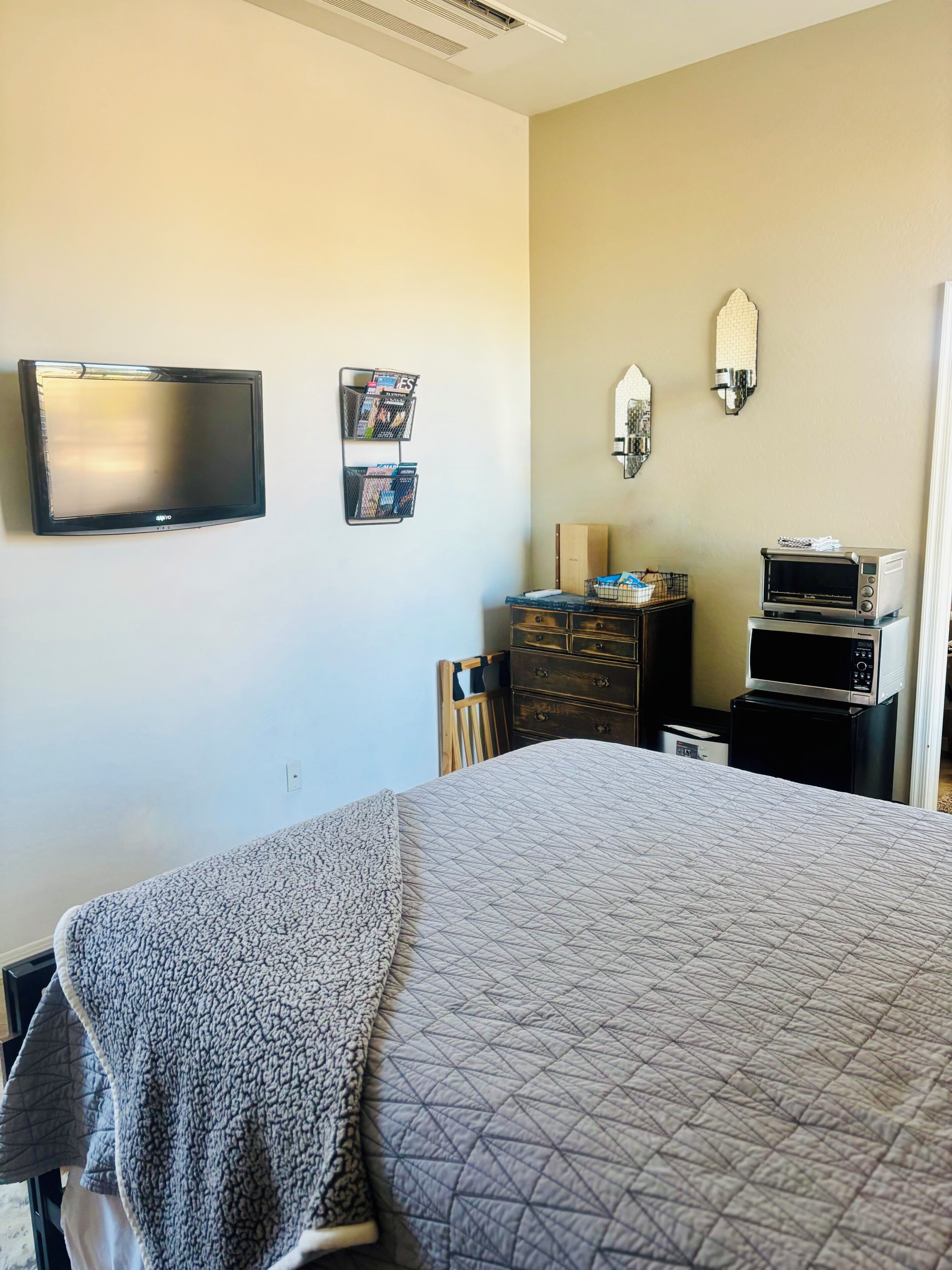 A hotel room with a bed, a flat-screen TV, a small kitchenette with a microwave and toaster, and a wall-mounted shelf with magazines.